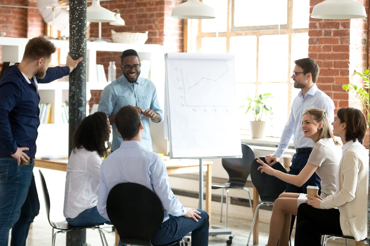 Smiling African American mentor or coach make flipchart presentation for diverse employees, excited black presenter present project on whiteboard, coworkers laugh having fun at casual briefing